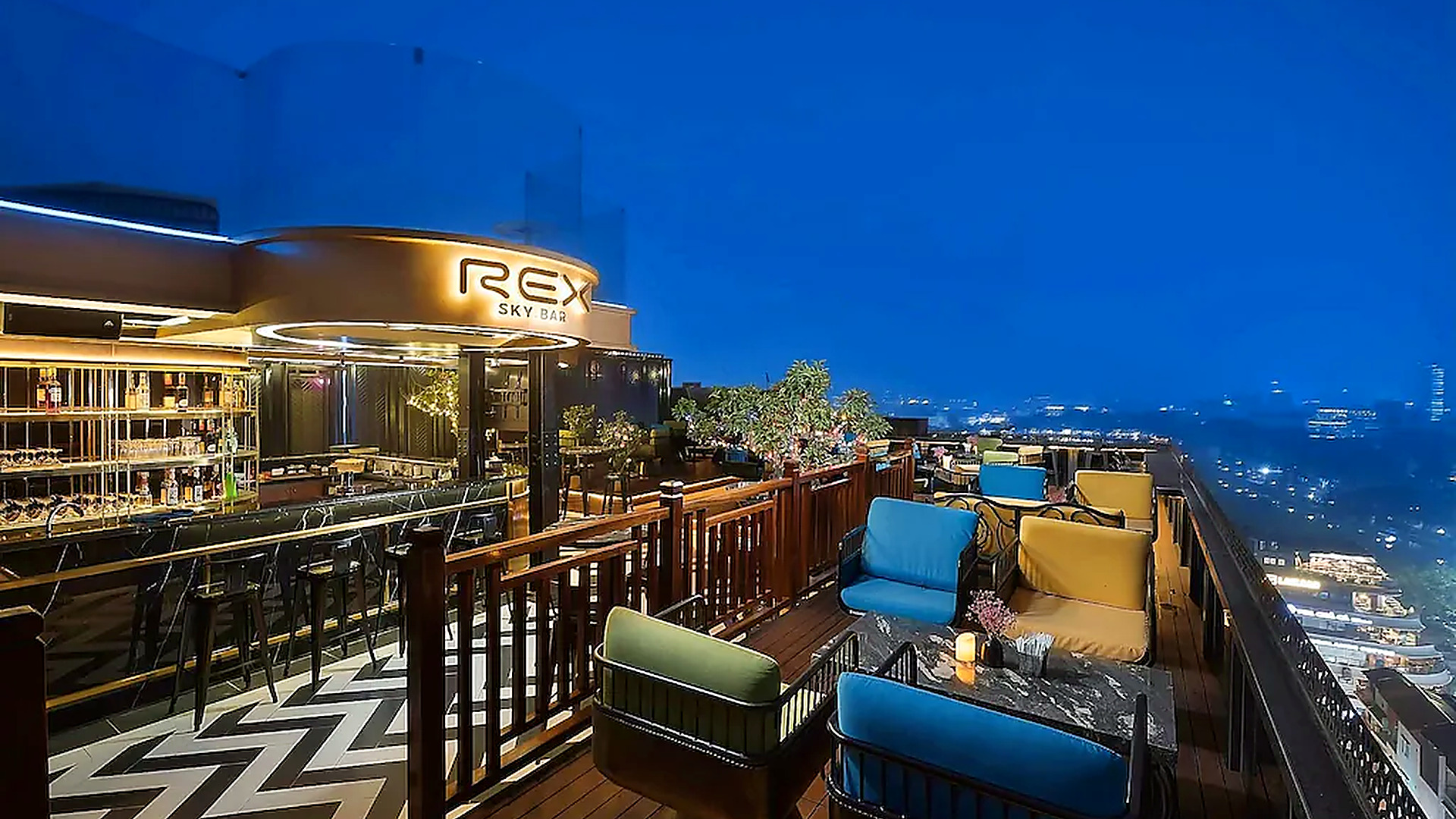 Sky bar rooftop về đêm với nhiều nhóm ghế ngồi, quầy bar sáng và tầm nhìn rộng ra thành phố.