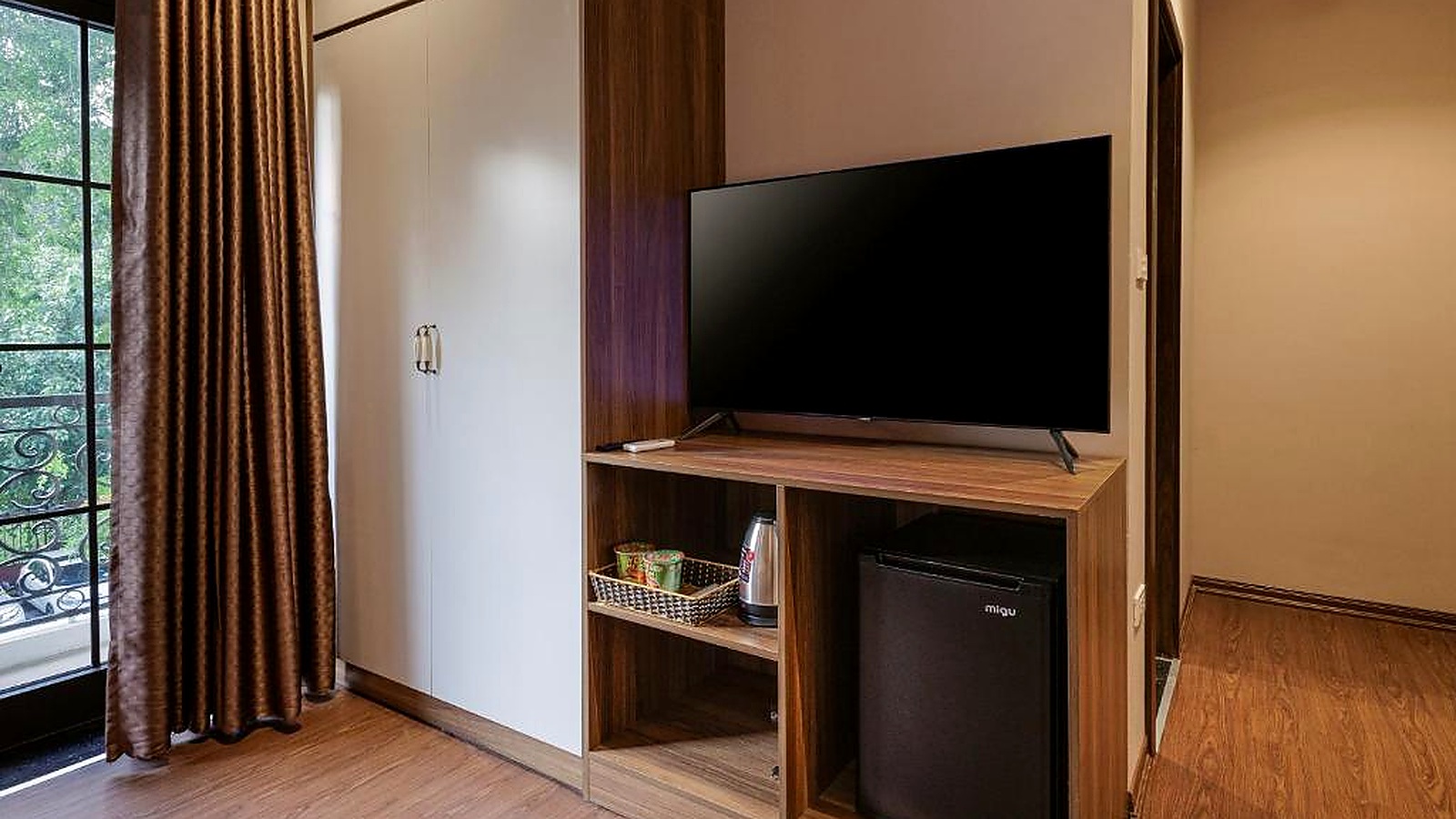 Khu tiện nghi phòng khách sạn gồm TV, tủ lạnh mini, khay đồ uống và tủ áo, được tích hợp gọn trong hệ nội thất gỗ.