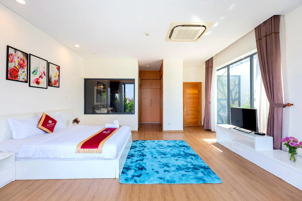 Phòng ngủ dùng nền trắng, sàn gỗ và cửa kính lớn; thảm xanh đóng vai trò điểm nhấn thị giác.