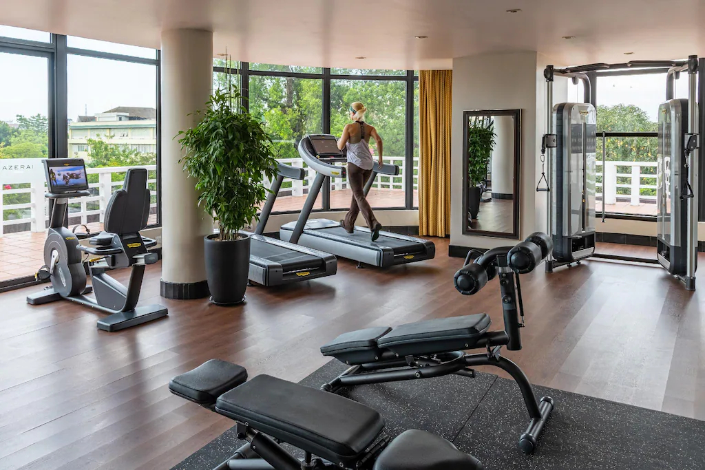 Gym thông thoáng dùng sàn gỗ, cây xanh và cửa rộng để giảm cảm giác khép kín của thiết bị tập.