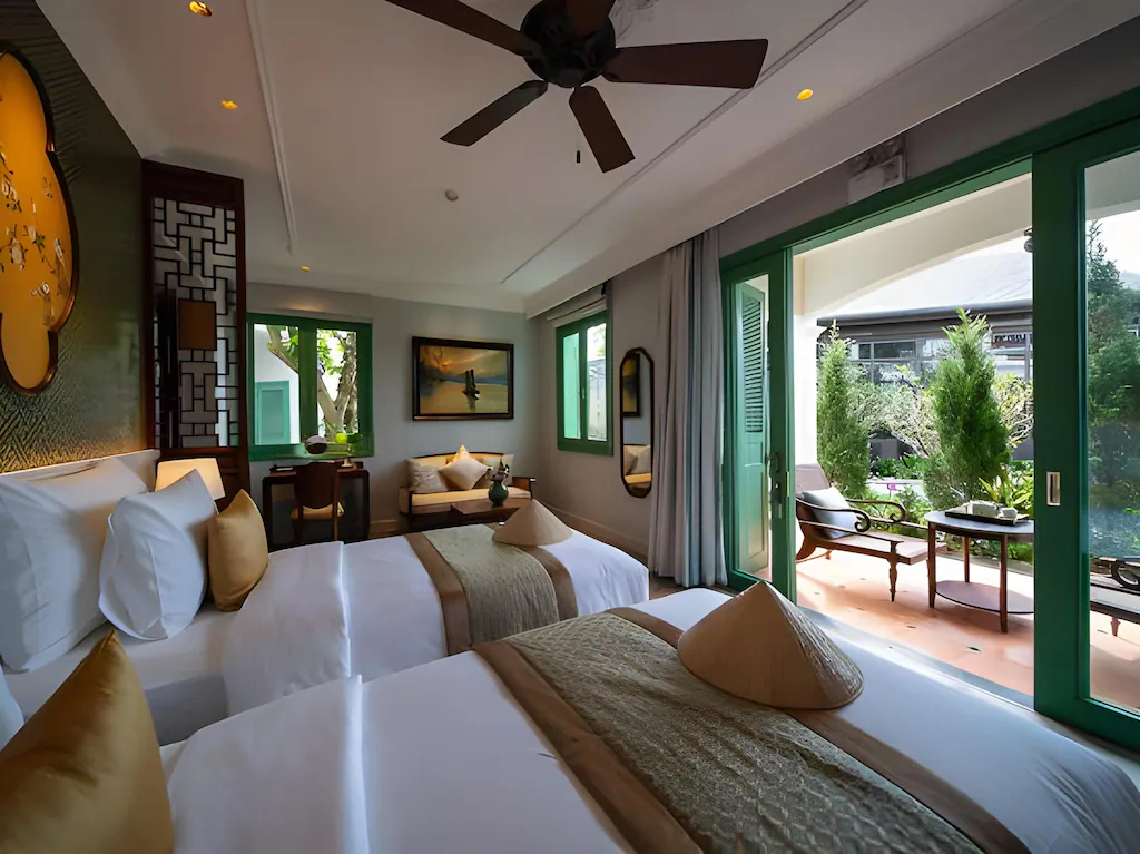 Hai giường lớn, khung cửa xanh và góc ngồi hướng vườn tạo hạng phòng linh hoạt cho gia đình hoặc bạn đồng hành.
