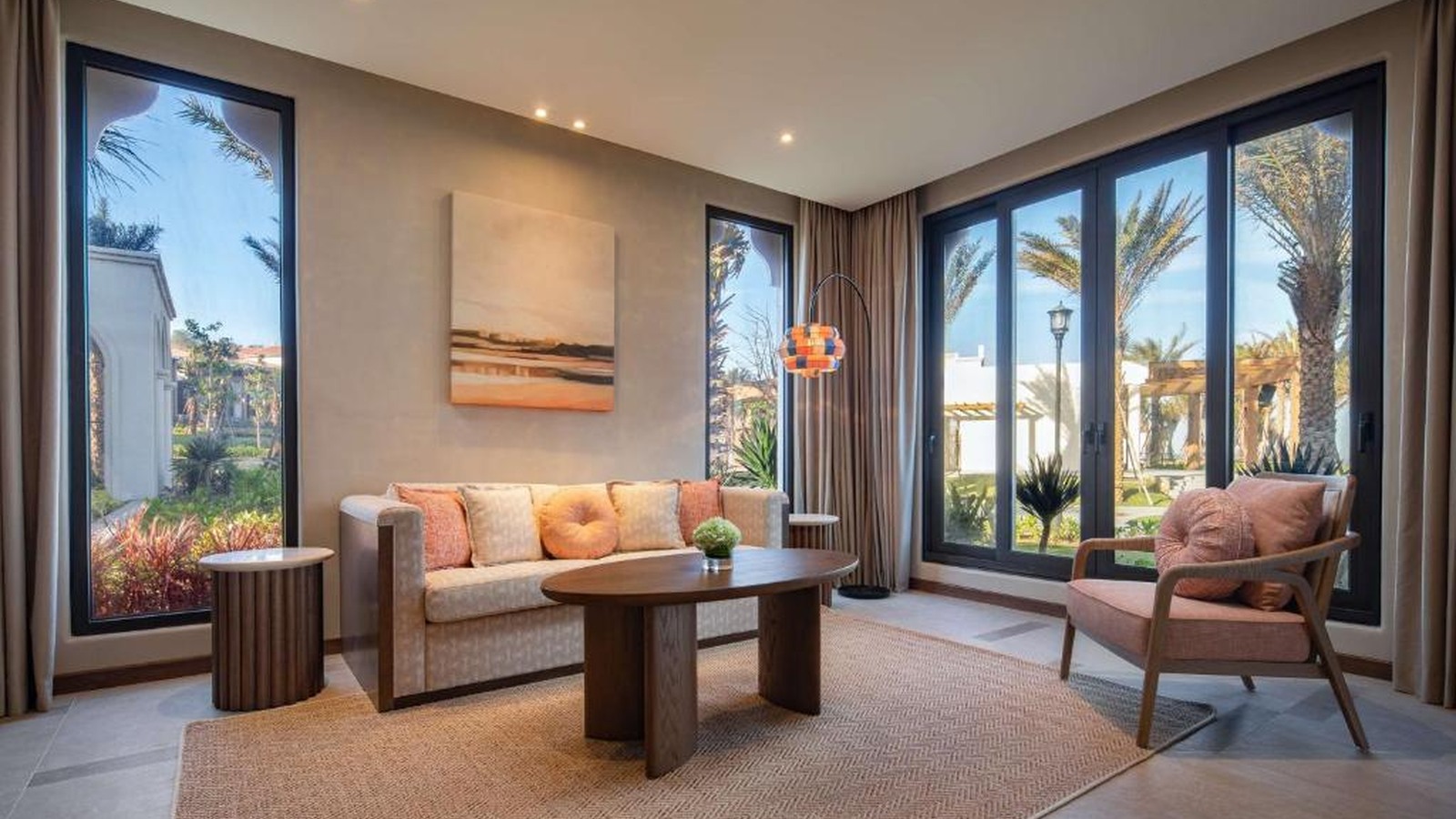 Phòng khách suite dùng sofa màu cát, ghế mây và cửa kính mở ra khu vườn nhiệt đới.
