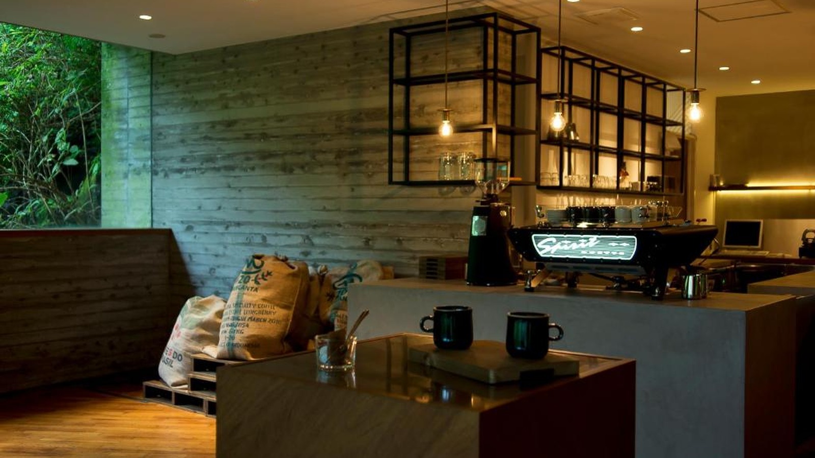 Quầy cafe nhỏ dùng gỗ, ánh sáng thấp và khung cửa xanh để tạo điểm dừng thân mật.