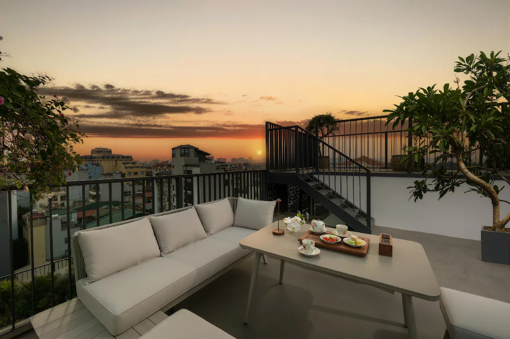 Lounge sân thượng khách sạn với sofa sáng màu, bàn trà và cảnh hoàng hôn thành phố