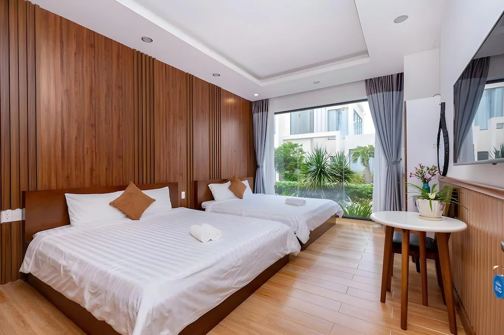 Phòng twin lặp lại ngôn ngữ gỗ, rèm xám và cửa kính nhìn vườn trong một bố cục dễ sử dụng.