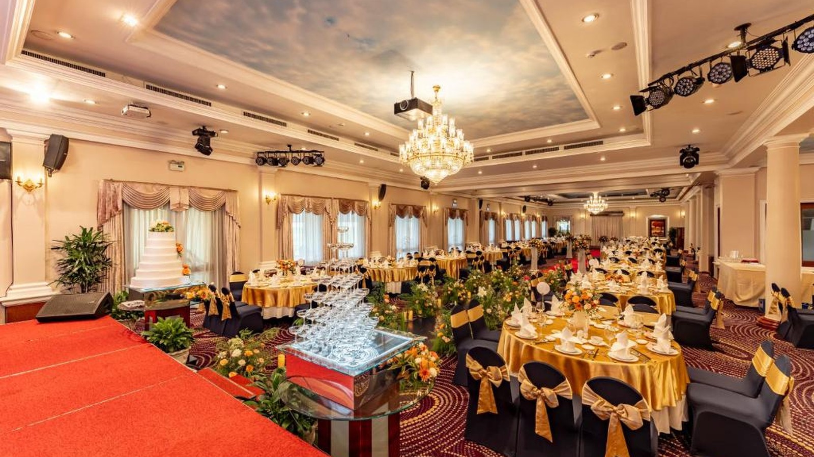 Ballroom sẵn sàng cho tiệc cưới với sân khấu, bàn tiệc tròn, hoa tươi và hệ đèn chùm
