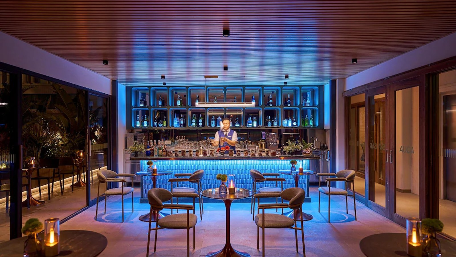 Bar lounge tạo không khí về đêm bằng ánh xanh, trần gỗ và các điểm sáng ấm trên bàn.