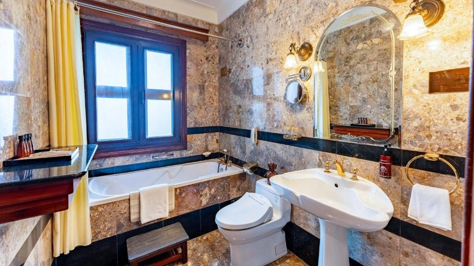 Phòng tắm ốp đá kết hợp bồn ngâm, gương vòm và chi tiết đồng để giữ nét Đông Dương sang trọng.